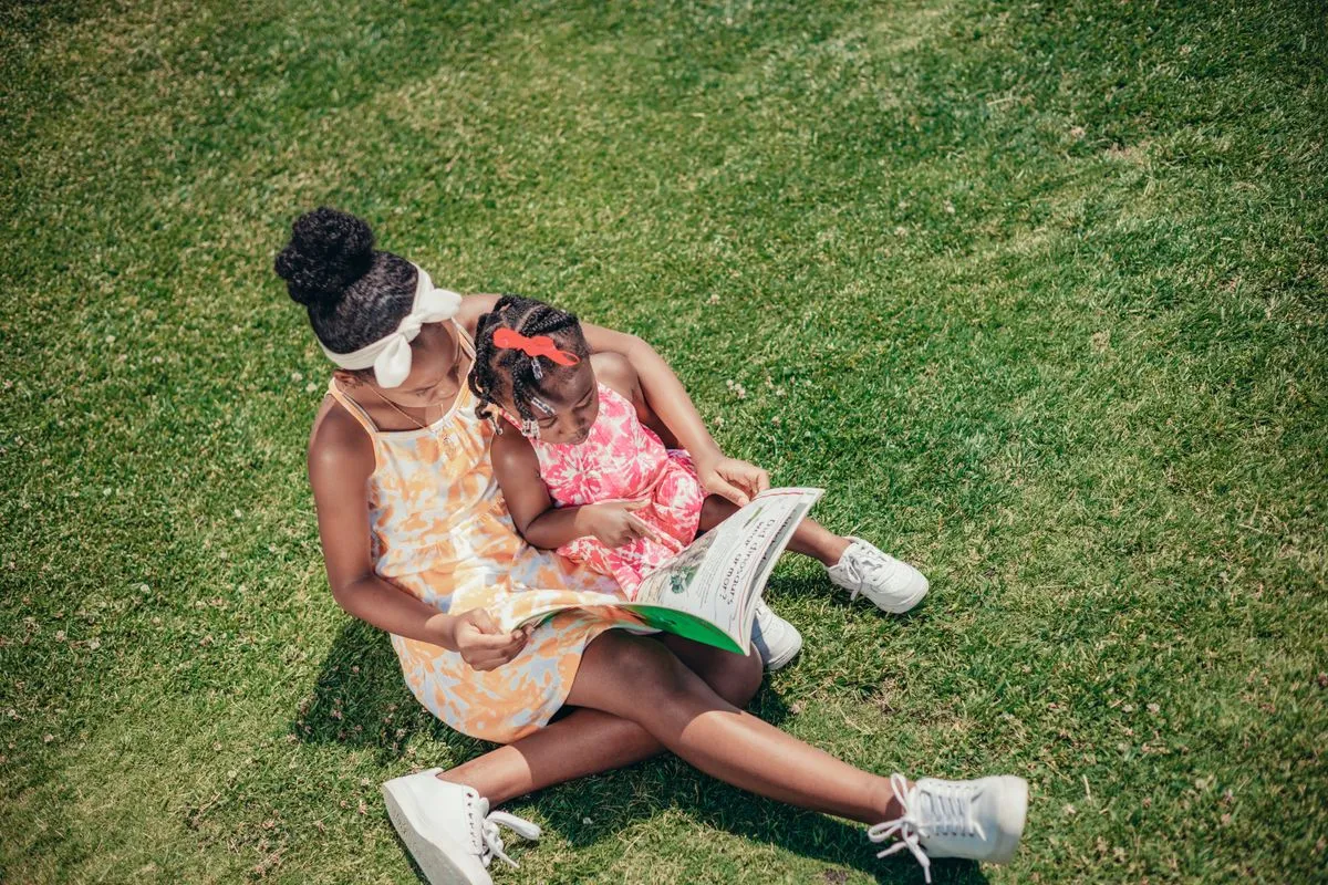 Two sisters reading a book together on the grass in a sunny park